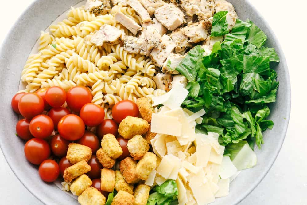 Chicken Caesar Pasta salad is fun twist on the classic! Fresh romaine tossed with chicken, cherry tomatoes, shredded parmesan cheese, croutons and a vegetable pasta adds a fun new texture! All the ingredients for Chicken Pasta Caesar Salad ready to be mixed.