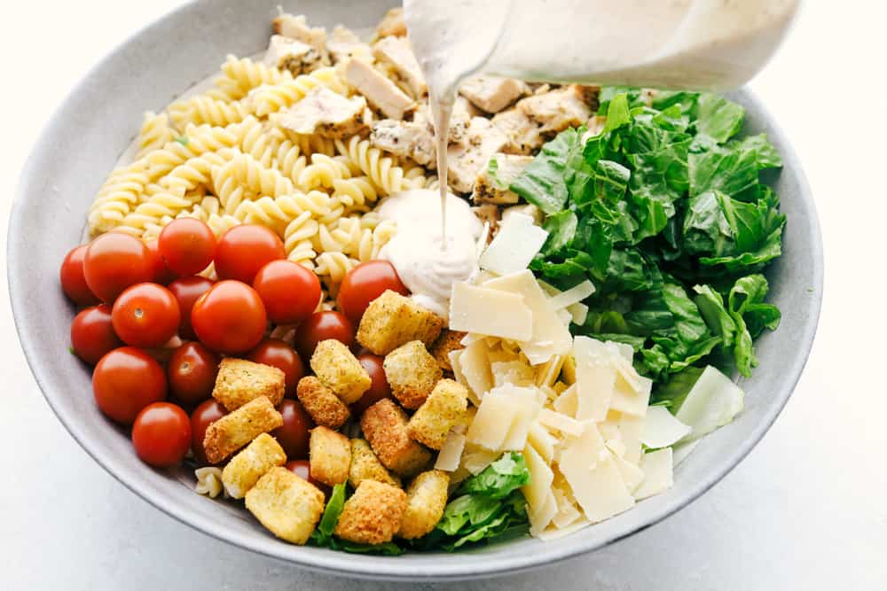 Chicken Caesar Pasta salad is fun twist on the classic! Fresh romaine tossed with chicken, cherry tomatoes, shredded parmesan cheese, croutons and a vegetable pasta adds a fun new texture! Pouring the dressing on the salad.