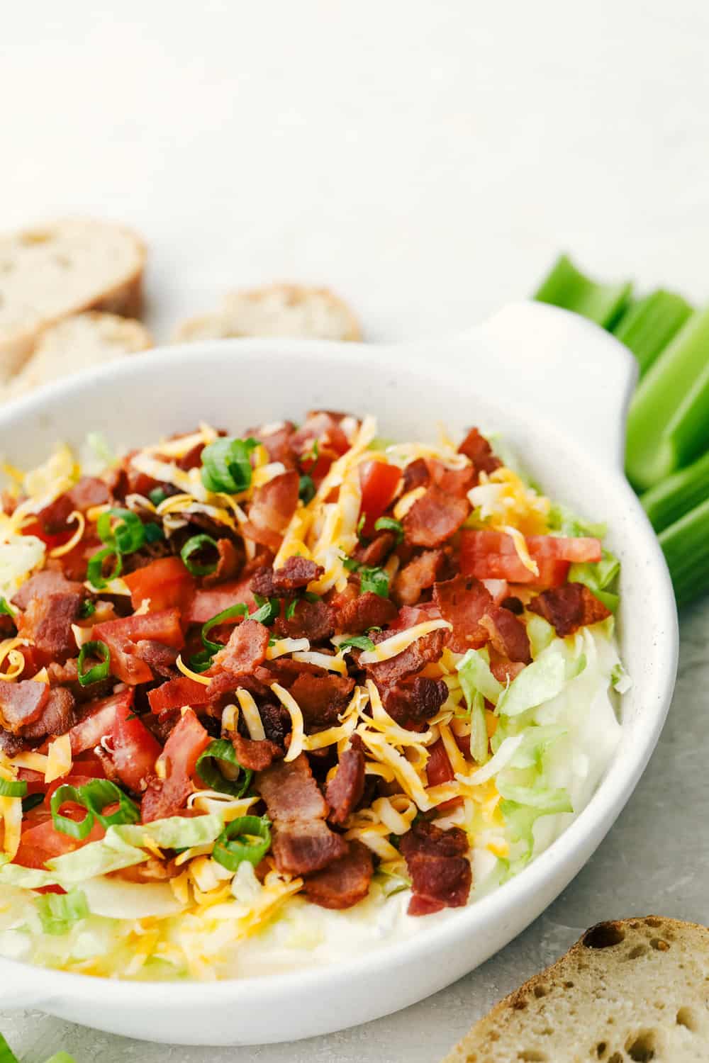 Creamy BLT Dip is an appetizer that is delightfully light and oh so good! A creamy seasoned base is topped with all the flavors of the classic BLT, you are going to love it! The finished BLT dip in a white bowl with options for dipping.