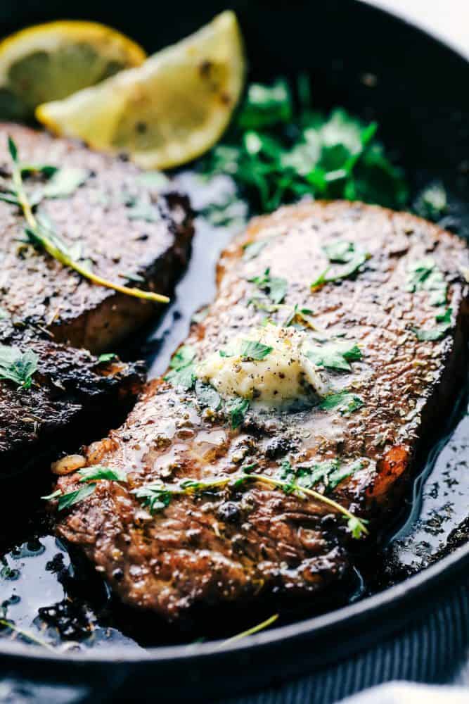 World's Best Steak Marinade adds so much flavor to your juicy steak. It helps to tenderize and infuse the steak with flavor and creates the best steak ever! Cooked steak in a skillet with marinade, lemons and garnish on top.