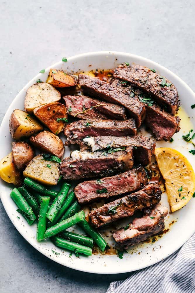 World's Best Steak Marinade adds so much flavor to your juicy steak. It helps to tenderize and infuse the steak with flavor and creates the best steak ever! Marinated and cooked steak with cooked potatoes, green beans and sliced lemons on a white plate.