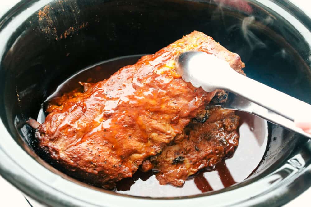 These Slow Cooker ribs are going to melt in your mouth and fall off the bone! They are cooked low and slow so they are perfectly tender and then covered in a sauce that's absolutely to die for! Removing the cooked ribs from the slow cooker.