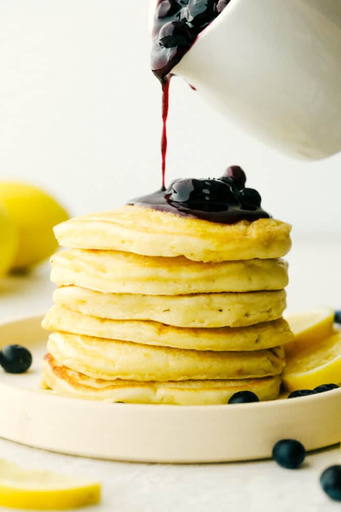 Lemon Ricotta Pancakes are fluffy, tender pancakes that are elevated to a whole new level. The ricotta adds protein and a lightness that is absolutely divine while the lemon gives it a bit of a tangy taste that you are going to love! Pouring blueberry sauce over a stack of lemon pancakes.
