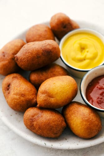 A crispy cornbread outer layer and snack-able size make these Mini Corn Dogs the perfect family treat! Recreate delicious county fair flavors in just a few simple steps! Mini corn dogs on a plate with mustard and ketchup on the side.
