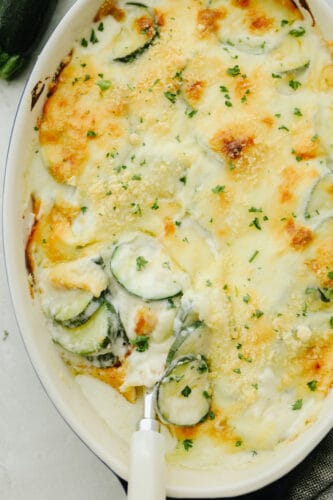 Three Cheese Zucchini Gratin has layers of creamy, cheesy sauce and is topped with a crunchy buttery topping. Ooey-gooey and cheesy, this low-carb take on comfort food will please any crowd! Parmesan Zucchini Gratin in a white dish.