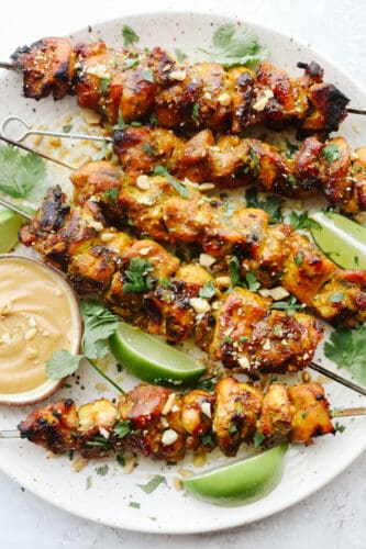 Chicken Satay is juicy, tangy, and absolutely divine. Marinated and then grilled to perfection serve these with a savory peanut sauce that is positively addicting. Grilled Chicken satay with peanut sauce on a plate.