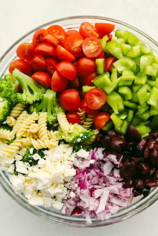 Greek Broccoli Pasta Salad combines tender fusilli pasta with fresh vegetables and a tangy herb dressing. This is the perfect dish to serve with grilled meats, or pack for a picnic or barbecue! A variety of vegetables and cheese in a bowl, ready to be mixed.
