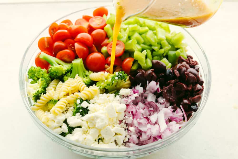 Greek Broccoli Pasta Salad combines tender fusilli pasta with fresh vegetables and a tangy herb dressing. This is the perfect dish to serve with grilled meats, or pack for a picnic or barbecue! Pouring dressing over pasta salad.