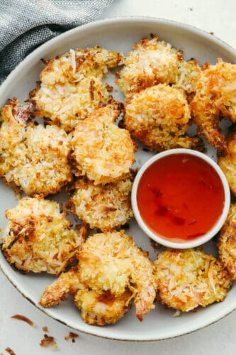 These super easy baked coconut shrimp will deliver in a flavor so delicious they will be sure to be a favorite among all of your guests! They are golden and crispy and baked to perfection. Baked Coconut Shrimp on a dish with serving sauce.