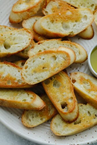 This easy, baked crostini is the best side to any Italian meal! They're golden, delicate pieces of heaven ready for your favorite toppings! Closeup of crostini pieces on a white plate.