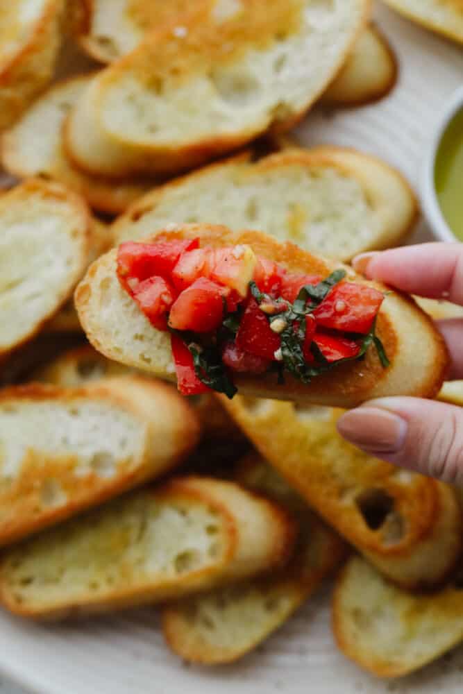 This easy, baked crostini is the best side to any Italian meal! They're golden, delicate pieces of heaven ready for your favorite toppings! Holding a piece of crostini topped with tomato basil salad.