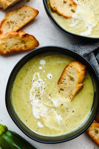 This zucchini soup has an amazing savory flavor, fresh vegetables, and is topped with the perfect drizzle of cream. Healthy and hearty, each spoonful will have you hooked with its silky-smooth goodness! 2 bowls of zucchini soup drizzled with cream.