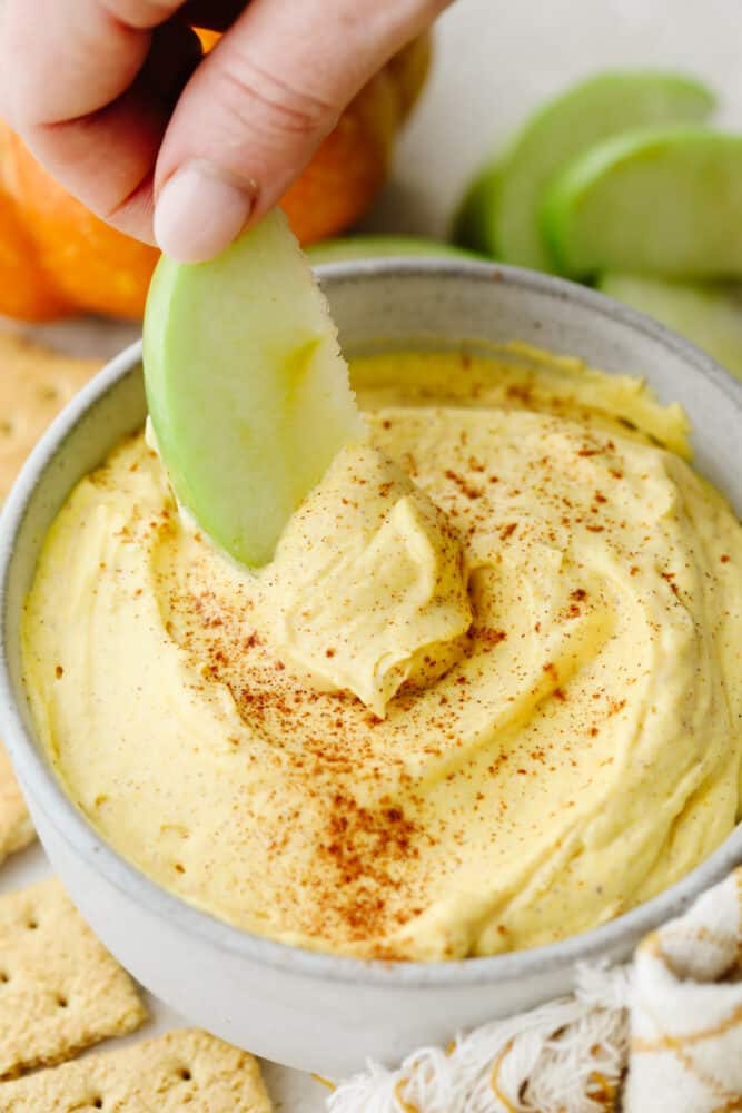 Pumpkin pie dip is deliciously light and fluffy! It has to creamiest texture and the spices bring that pumpkin pie flavor to life! It is something that your whole family will go crazy over! Someone dipping an apple into pumpkin pie dip.