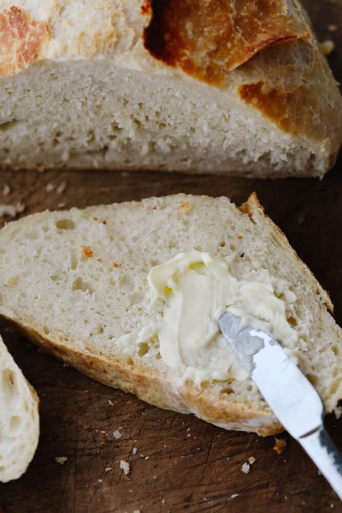 This amazing artisan no-knead bread recipe will make you feel like a rock-star baker! It has a perfectly crispy crust with a soft and chewy texture on the inside. Plus, it couldn't be easier to make from home! A knife spreading butter onto a slice of bread.