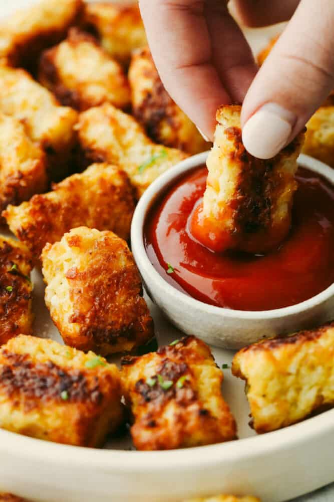 Cauliflower tots are healthy and delicious! Baked to absolute perfection, these tots are crispy on the outside and soft on the inside. Everyone will love them so much that they will fly off the table! A cauliflower tot being dipped into ketchup.