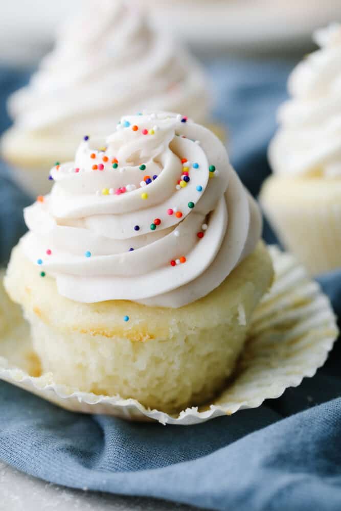 White cupcakes are light, fluffy and perfectly moist! Swirled with delicious white frosting and topped with sprinkles, these cupcakes are to die for! A white cupcake with the liner pulled off.