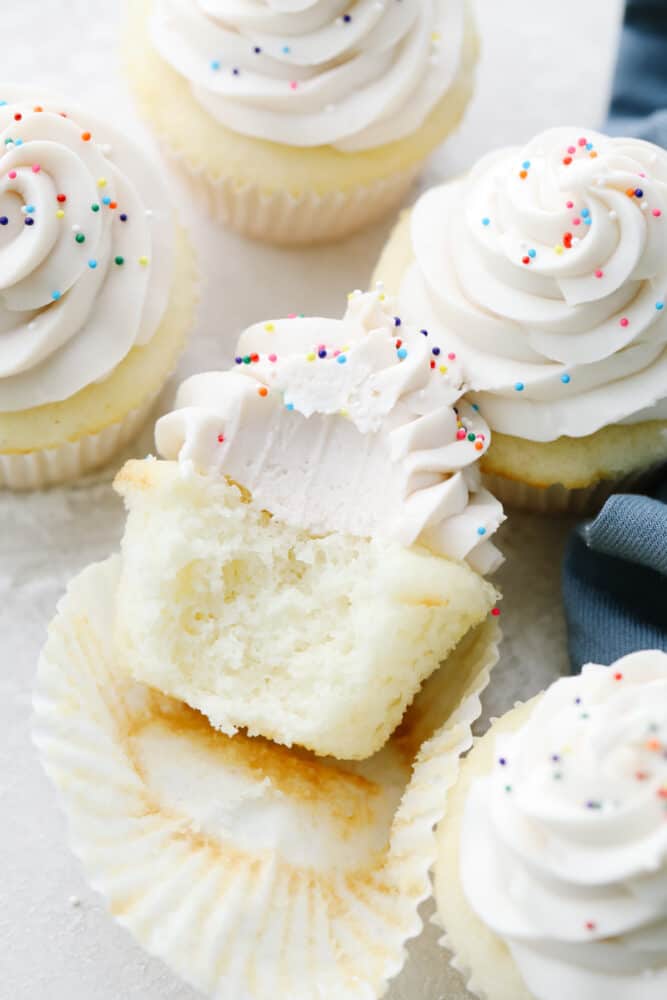White cupcakes are light, fluffy and perfectly moist! Swirled with delicious white frosting and topped with sprinkles, these cupcakes are to die for! A white cupcake with a bite taken out of it.