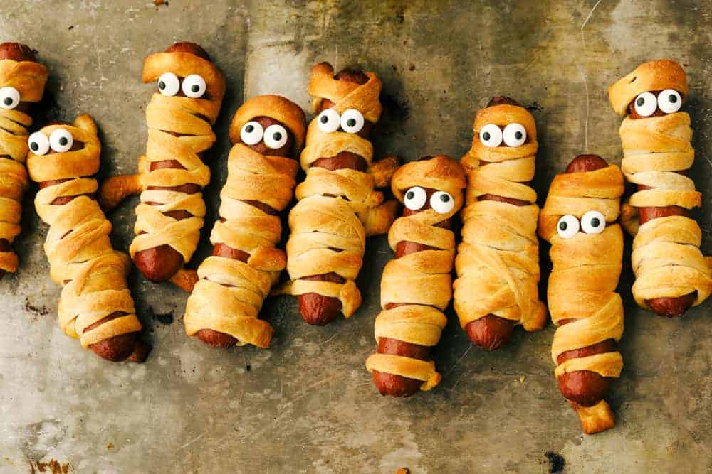 Nothing screams fun, quite like Mummy Hot Dogs for Halloween dinner. Serve these fun little vittles before trick or treating or at your next Halloween party. Mummy dogs lines up on a tray.
