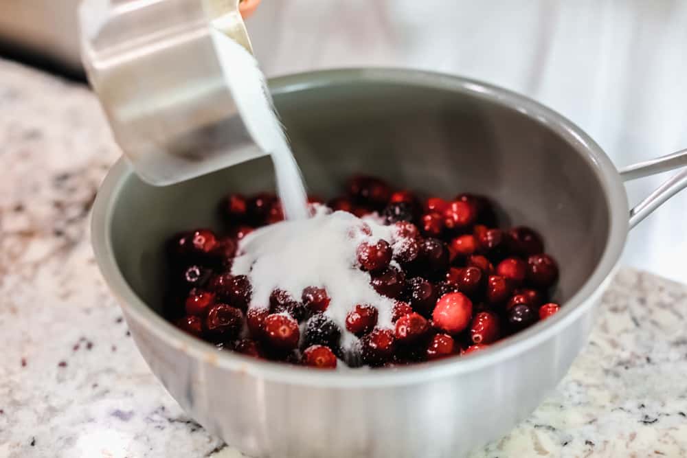 This cranberry sauce is the perfect mixture of sweet and tart! It's made with fresh cranberries and topped with orange zest to really set it apart! Sugar being poured onto cranberries.