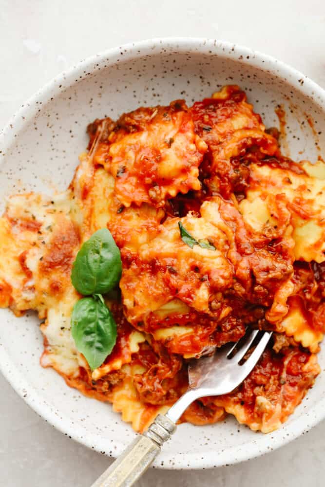 This baked ravioli is a knockout dinner that is sure to be loved by all! It's smothered in melty mozzarella, parmesan, and a tasty meat sauce! Baked ravioli in a white stoneware bowl.