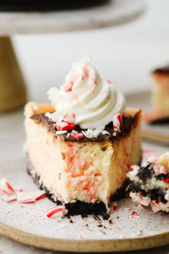 Peppermint cheesecake is creamy and delicious! It has the perfect oreo crust and is topped with crushed peppermint and whipped cream. This will definitely get a "WOW!" from all of your guests this holiday season! A slice of peppermint cheesecake on a plate with a bite taken out of it.