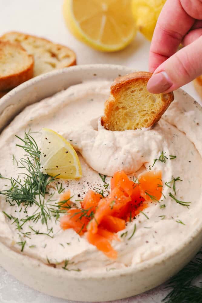 This smoked salmon dip is so creamy and delicious! With the smoked salmon flavor taking the stage followed by a hint of lemon and dill, this dip is SOO GOOD! A crostini slice being dipped into smoked salmon dip.