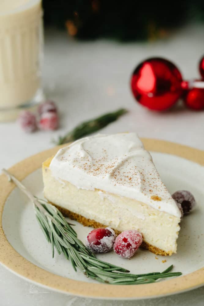 This eggnog cheesecake is SOO GOOD. It's creamy and melts in your mouth! That classic eggnog taste makes it the best dessert for the holiday season! A slice of cheesecake on a plate with sugared cranberries.
