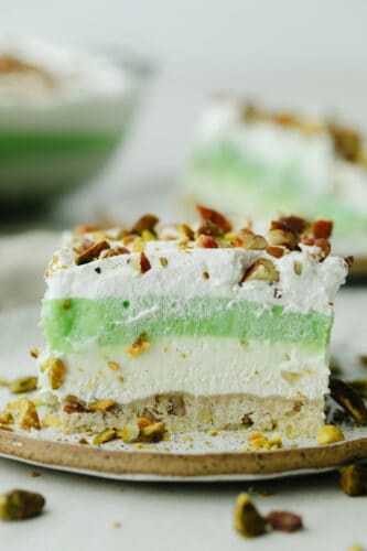 This layered pistachio dessert is insanely delicious! It has the perfect crust, topped with layers of creamy cheesecake and pistachio pudding. The crushed pistachios on the top finish it off and make it the best dessert for any occasion! A slice of layered pistachio dessert on a plate.