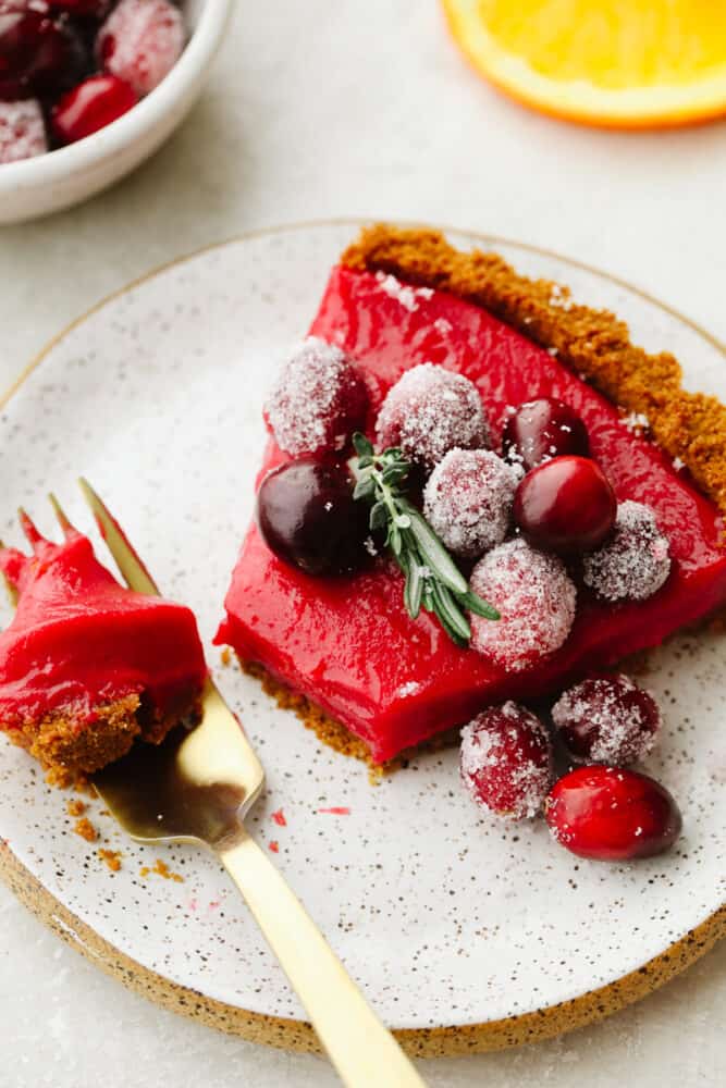 With a smooth tangy filling and Biscoff crust, this cranberry curd tart is a must-make for the holidays. It's as delicious as it is beautiful! A slice of cranberry curd tart on a plate with a fork bite.
