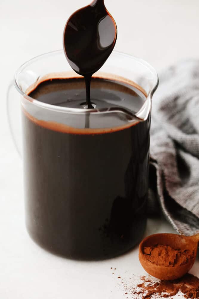 This homemade chocolate sauce has the perfect chocolate flavor and the smooth texture is to die for. It goes on just about anything and is so simple to make! A spoon being pulled out of a pitcher filled with chocolate sauce.