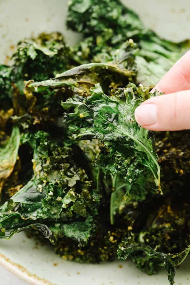 Crispy and seasoned perfectly with parmesan cheese, these kale chips that you cook in the air fryer are the BEST snack. They are quick and easy to make and I bet that you can't stop at just one! A hand picking up a kale chip to eat.