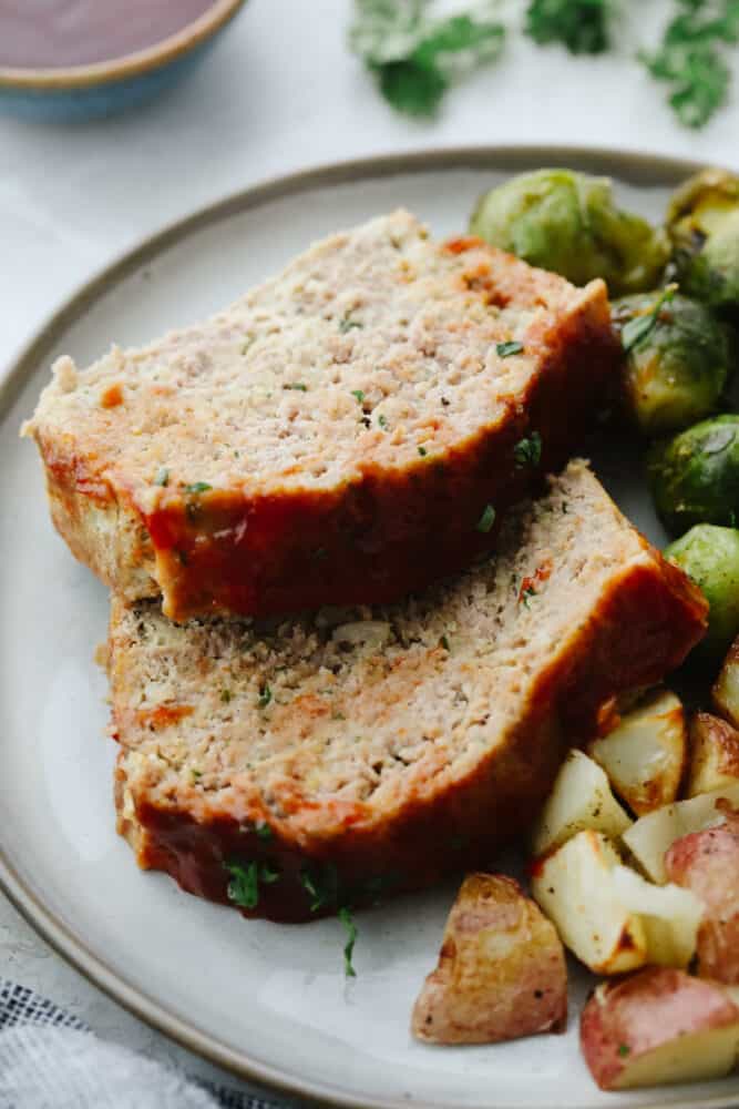 This tasty turkey meatloaf is light and lean but filled with so much flavor! Each bite is perfectly seasoned and tender, you'll love it! 2 slices of turkey meatloaf on a plate with potatoes and brussels sprouts.