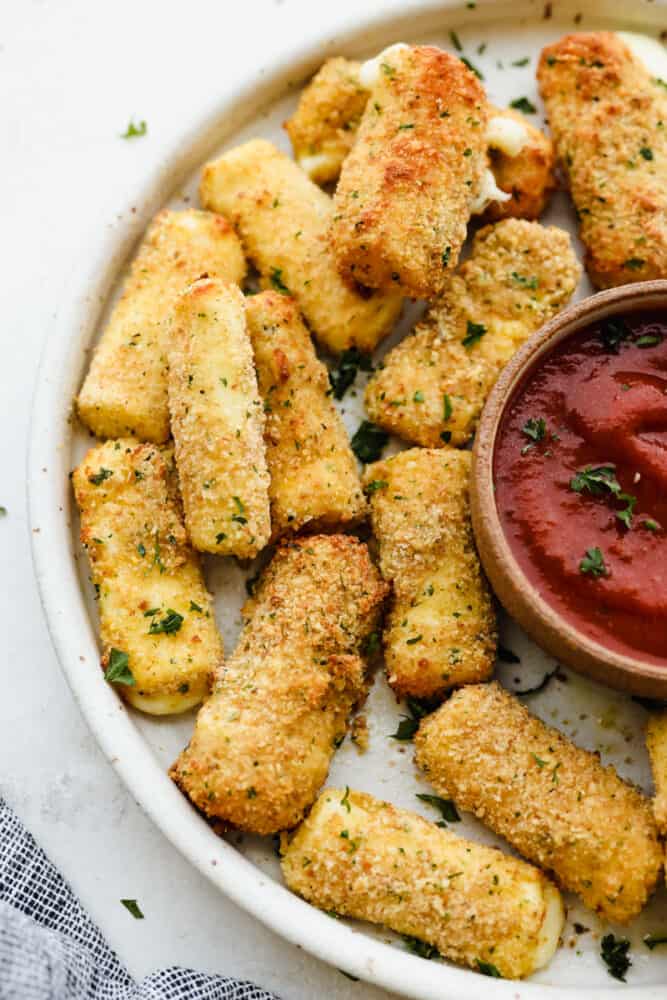 Crispy, cheesy, and delicious, these easy air fryer mozzarella sticks are going to be your new favorite snack! They're super simple to prepare and cook up in less than 10 minutes! Air fryer mozzarella sticks on a white plate served with marinara sauce.
