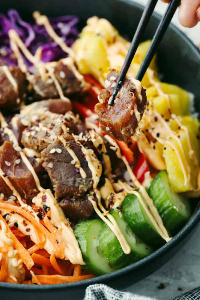This Hawaiian poke bowl is fantastically fresh and full of healthy ingredients! Skip ordering this at a restaurant and make it at home in less than 15 minutes! A hand holding chopsticks with a piece of tuna coming out of a Hawaiian poke bowl