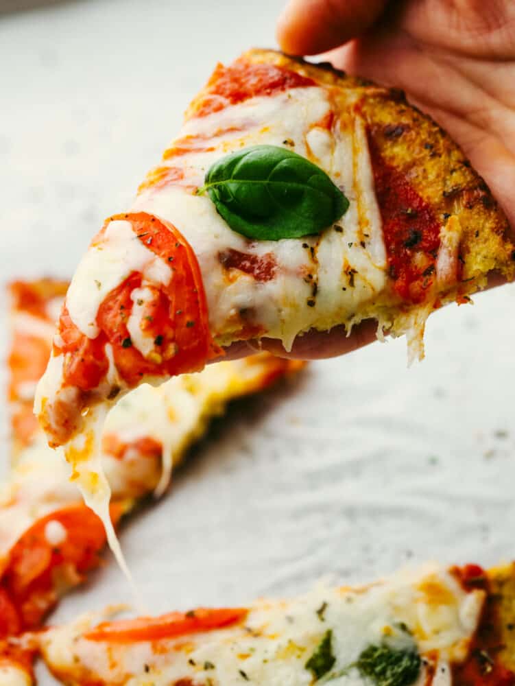 This cauliflower pizza crust is grain-free and absolutely delicious!! It's so easy to make and when you are done, you won't be able to even tell that it's the healthy version! It's THAT good! A slice of pizza being held in a hand.
