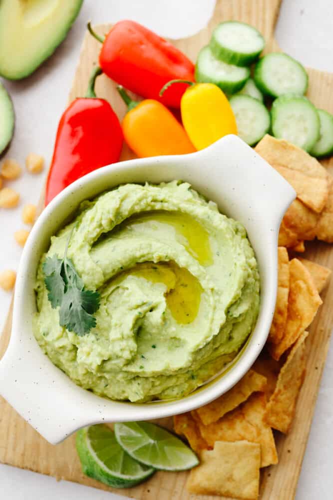 If you are looking for the most delicious appetizer out there, this avocado hummus is for you! Delectable, creamy, and perfect with any veggies or crackers, this dip is the BEST! Avocado hummus in a bowl on a wooden cutting board with some pita chips, cucumbers and peppers.