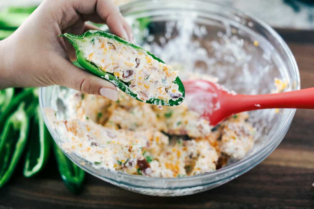 These jalapeño poppers are filled with a cream cheese mixture, topped with crumbled bacon pieces, and then baked to perfection. You won't be able to stop at one! Jalapeño poppers being stuffed with cream cheese, cheese and bacon mixture.
