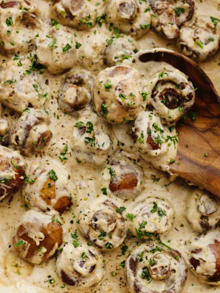 Calling all mushroom lovers! These creamy garlic parmesan mushrooms are waiting for you! Sautéed in garlic butter until tender and then tossed in the most AMAZING creamy parmesan sauce, these easy mushrooms will be off the plate and in your mouth in less than 10 minutes! Creamy, garlic parmesan mushrooms being served with a wooden spoon.