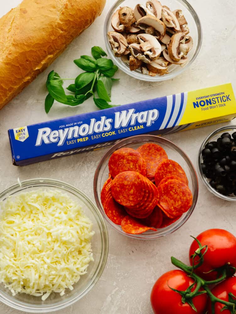These foil french bread pizzas made with Reynolds Wrap® Non-Stick Foil are perfect for any family dinner! Crispy, cheesy, and delicious, your family won't be able to resist coming back for seconds! Reynolds Wrap® Non-Stick foil, pepperonis, mushrooms, tomatoes, basil, olives, and cheese in bowls on the counter.