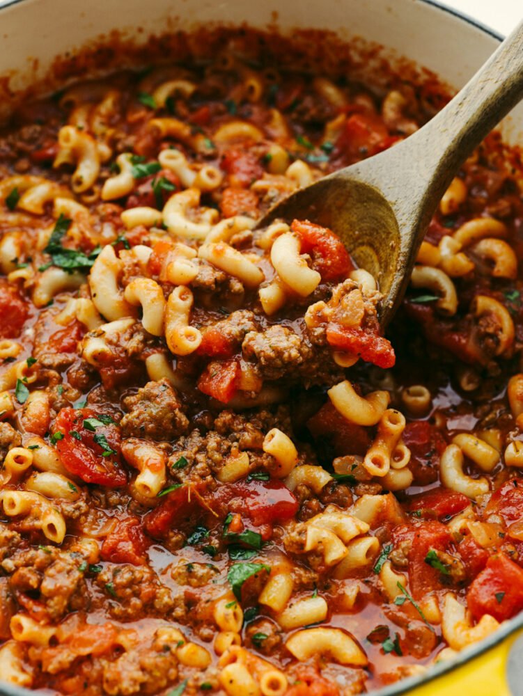 American Goulash is an easy old-fashioned comforting meal that is perfect for the cold winter months. Everything cooks in the same pot, even the pasta! A pot with American goulash being stirred with a wooden spoon.