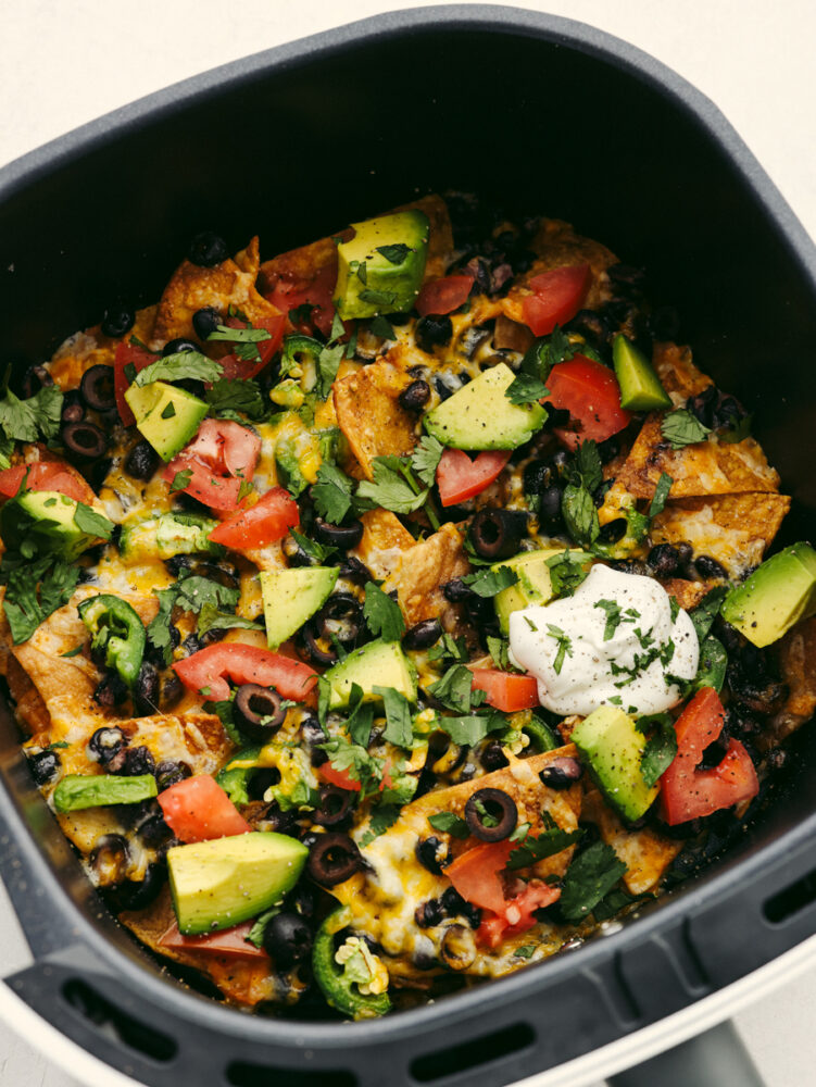 Air fryer nachos are loaded with cheese and vegetables and cooked to absolute perfection. This quick and easy appetizer is perfect for game day or any family get-together! An air fryer loaded with nachos topped with cheese, vegetables, sour cream and cilantro.