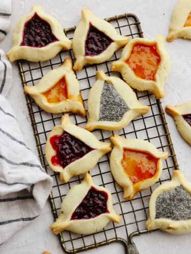 These triangle-shaped hamantaschen cookies are perfectly soft and filled with delicious jams and a homemade poppyseed filling. Step up your game and impress your guests with this unique and flavorful dessert! Hamantaschen on a baking rack cooked and ready to eat.
