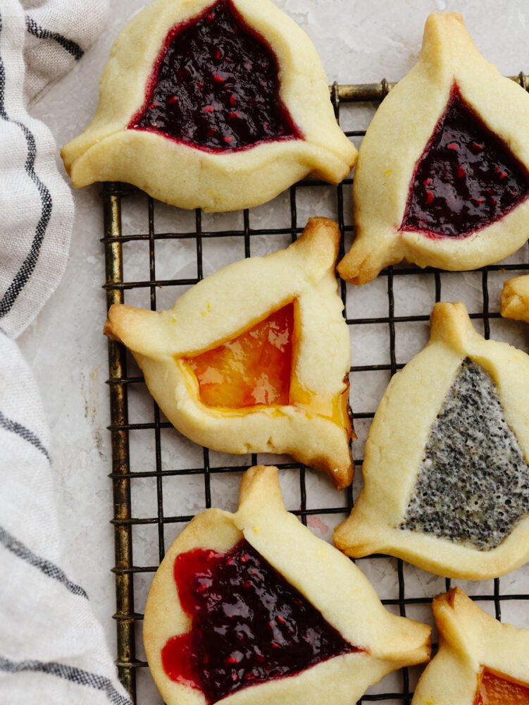 These triangle-shaped hamantaschen cookies are perfectly soft and filled with delicious jams and a homemade poppyseed filling. Step up your game and impress your guests with this unique and flavorful dessert! Hamantaschen on a cooling rack.