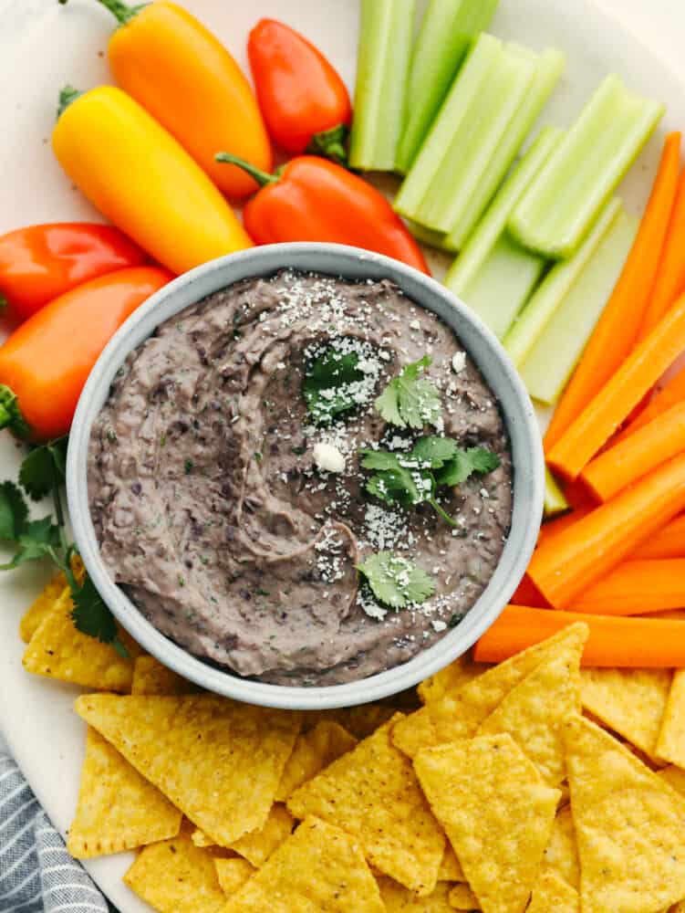 This black bean dip flavored with garlic, lime, and spices is going to be the star of your next party! It whips up in minutes and is absolutely irresistible. Black bean dip in a white bowl, surrounded by chips and vegetables.