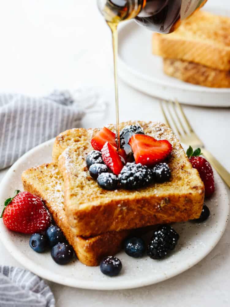 Air fryer french toast is simple and delicious! You can whip this up in less than 10 minutes, and your family will gobble it up so fast that it will fly off of your table! 2 pieces of french toast on a plate with berries and powdered sugar with syrup being poured on top.