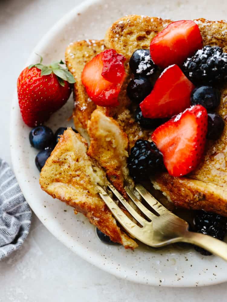 Air fryer french toast is simple and delicious! You can whip this up in less than 10 minutes, and your family will gobble it up so fast that it will fly off of your table! A piece of french toast on a plate with berries and syrup, being cut into with a gold fork.