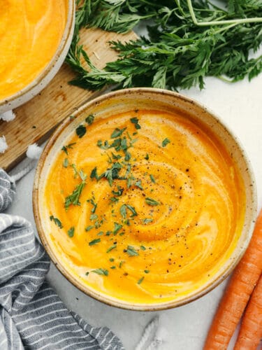 Carrot soup is the perfect combination of fresh and hearty. It's creamy, delicious, and flavored to perfection and your guests will be running back for seconds! A top view of a bowl of carrot soup next to some large carrots.