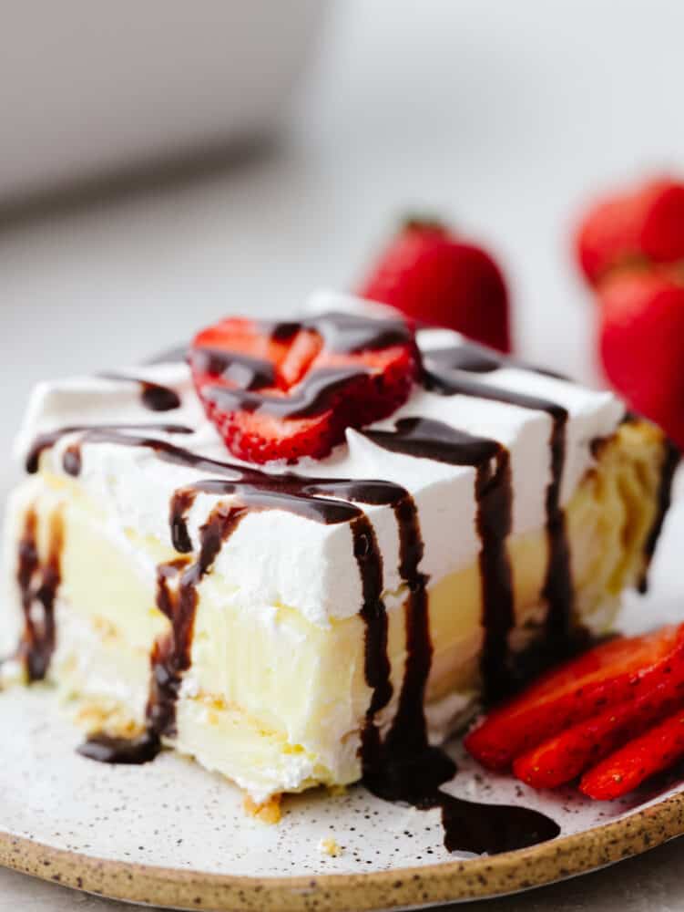 Eclair cake is moist and sweet, made with vanilla filling, a buttery crust, and whipped cream topping. There are so many toppings to choose from and you will be amazed at how insanely soft and delicious this cake is! A slice of eclair cake topped with chocolate drizzle and sliced strawberries.