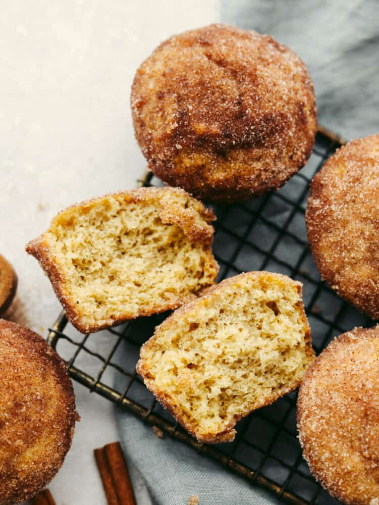Donut muffins are soft and fluffy, with a cinnamon-sugar flavored goodness that melts in your mouth! They are delicious enough for anyone to want to devour them the moment that you put them on the table! Donut muffins on a cooling rack with one broken in half so that you can see the inside.