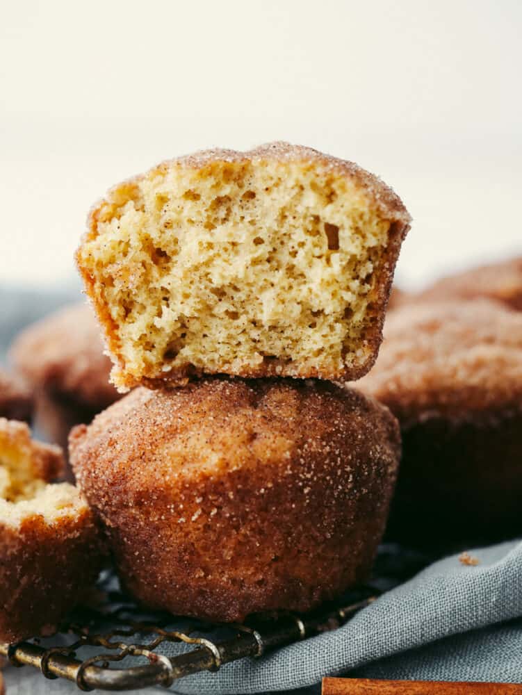 Donut muffins are soft and fluffy, with a cinnamon-sugar flavored goodness that melts in your mouth! They are delicious enough for anyone to want to devour them the moment that you put them on the table! 2 donut muffins stacked. The top on has a large bite taken out of it.
